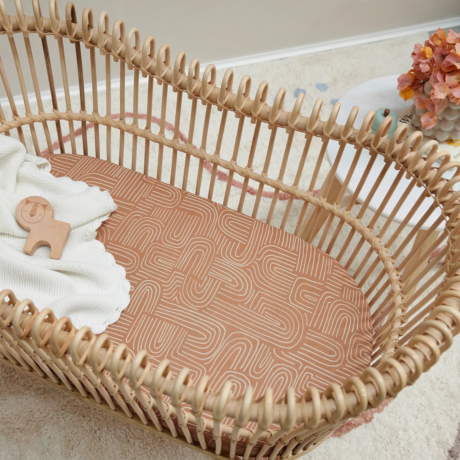 A natural rattan bassinet with the Mulberry Threads Bamboo Bassinet Sheet – Arches Design, a cream knit blanket, and a wooden toy on top. A white side table with pink flowers sits nearby on a light carpet.