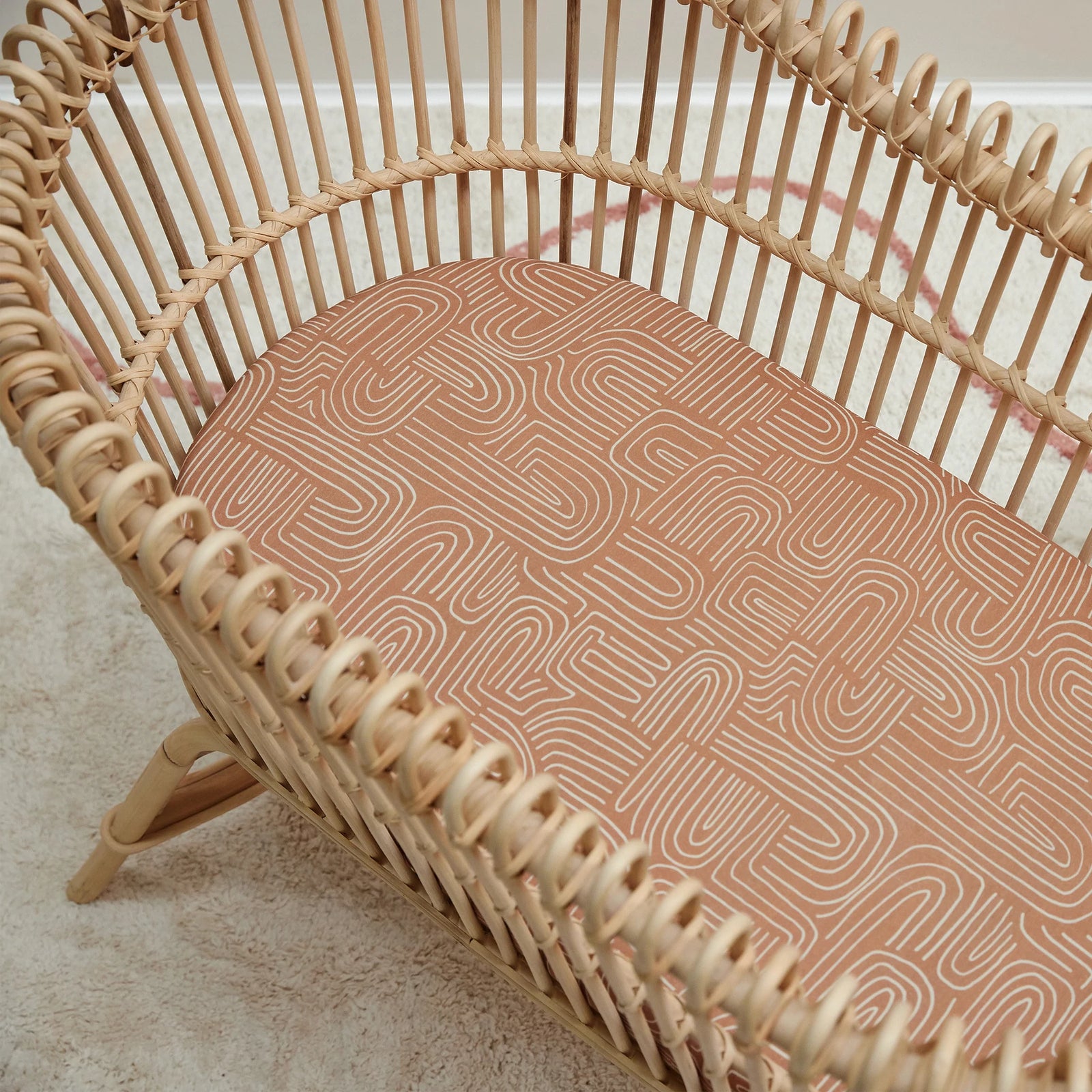 A natural rattan bassinet with the Mulberry Threads Bamboo Bassinet Sheet – Arches Design, a cream knit blanket, and a wooden toy on top. A white side table with pink flowers sits nearby on a light carpet.